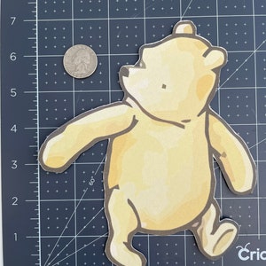 May include: A yellow Winnie the Pooh character cutout with a brown outline. The character is walking with its arms outstretched and is looking up.
