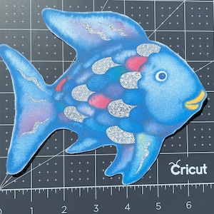 Pu&ograve; includere: Un pesce blu con squame di glitter argentate e una bocca gialla. Il pesce ha un design rosa e blu sul suo corpo. Il pesce &egrave; su uno sfondo a griglia bianco e nero. Il testo "Cricut" &egrave; visibile nell'angolo in basso a destra dell'immagine.