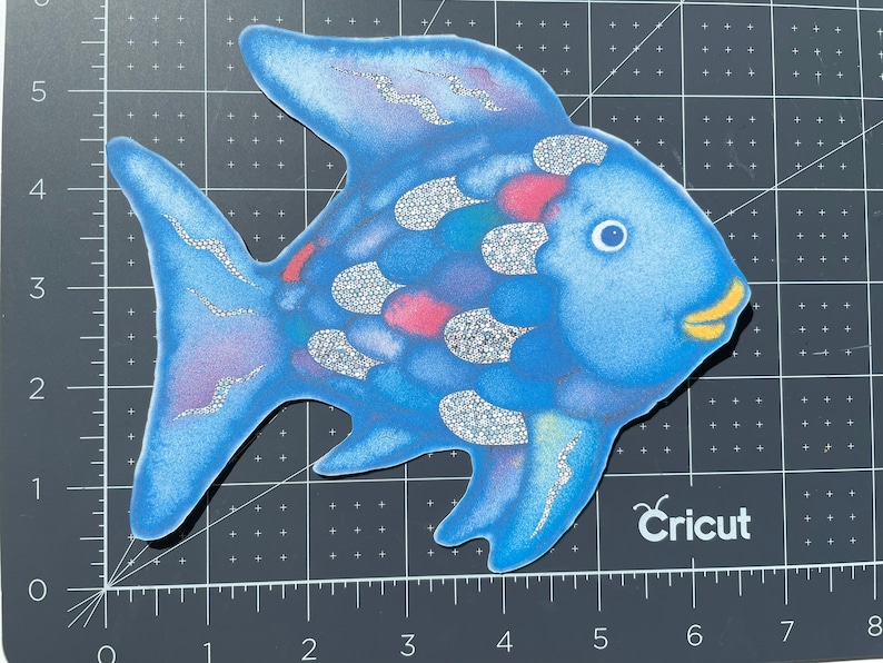 Pu&ograve; includere: Un pesce blu con squame di glitter argentate e una bocca gialla. Il pesce ha un design rosa e blu sul suo corpo. Il pesce &egrave; su uno sfondo a griglia bianco e nero. Il testo "Cricut" &egrave; visibile nell'angolo in basso a destra dell'immagine.