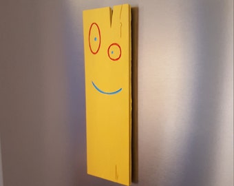 May include: A yellow wooden plank with a painted face. The face has two red circles for eyes with blue pupils and a blue smile.