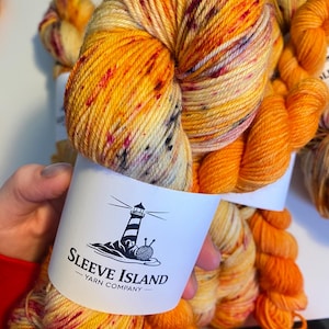May include: Close-up of vibrant orange and yellow yarn skeins with red and black speckles. The yarn is wrapped with a white label featuring a lighthouse and the text "Sleeve Island - Yarn Company".