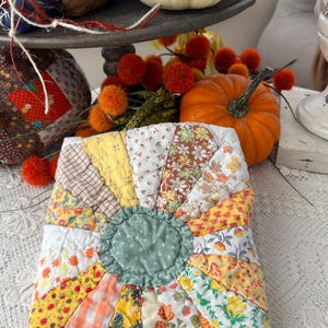 May include: A square quilted potholder featuring a central sage green circle surrounded by radiating fabric panels in various patterns including yellow, orange, white, and brown floral, gingham, and leaf prints with visible stitching.