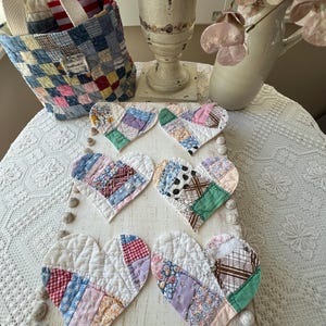 May include: A collection of six heart-shaped quilted coasters in various patchwork patterns, arranged on a distressed white wooden tray. A denim patchwork basket with a white handle and a vase with pink flowers are in the background.