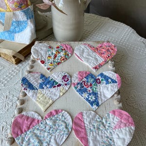 May include: Six heart-shaped quilted coasters in a variety of floral and solid fabrics. The coasters are arranged on a distressed white wooden tray. A white tote bag with blue and yellow patchwork is visible in the background.