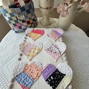 May include: A collection of six heart-shaped quilted coasters in various pastel colors, arranged on a distressed white wooden board. A patchwork tote bag and decorative vase are in the background, suggesting a vintage or farmhouse aesthetic.
