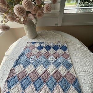 May include: A rectangular quilted textile featuring a diamond pattern composed of alternating blue, white, and muted mauve fabric squares. The quilt displays visible stitching throughout the surface, with some squares showcasing subtle grid or striped textures.