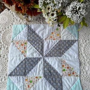May include: A square, handmade quilt block featuring a starburst design. The block is composed of white, gray, and patterned fabric triangles. The corners are light blue. The patterned fabric includes teapots and fruit. The quilt block is on a white lace surface.
