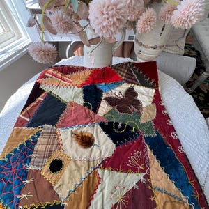 May include: A rectangular fabric wall hanging featuring a crazy quilt design with various patches in shades of red, brown, blue, and cream. The piece is decorated with hand-stitched embroidery, including a butterfly, a leaf, and spiderweb motifs.
