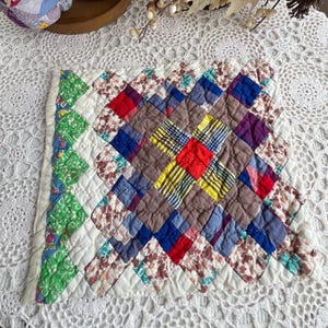 May include: A small, handmade quilt with a diamond pattern. The quilt features a variety of fabrics in colors like red, blue, yellow, and brown. The edges are finished with a white border and green patterned fabric.