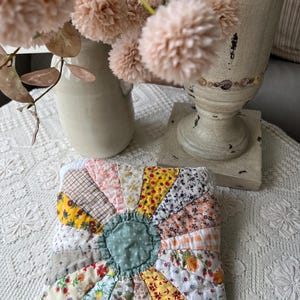 May include: A square quilted potholder featuring a central sage green circle surrounded by radiating fabric segments in various floral, gingham, and plaid patterns in shades of yellow, pink, white, and brown with visible stitched seams.