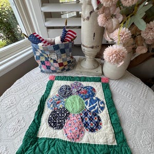 May include: A rectangular quilted wall hanging featuring a central flower motif with multi-colored patterned fabric petals and a solid light green center, framed by a cream-colored background and a wide, solid dark green border with pink tabs.