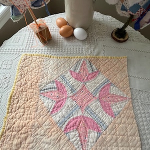 May include: A vintage quilt with a central diamond pattern in pink and white, set against a peach-colored background. The quilt features intricate stitching and a yellow border, showcasing a classic patchwork design. The quilt is displayed on a white lace tablecloth.