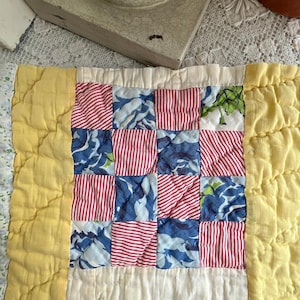 May include: A handmade quilt with a patchwork design. The quilt features squares of red and white striped fabric, blue floral fabric, and green floral fabric. The quilt is bordered with yellow fabric and has a quilted texture.