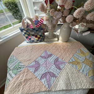 May include: A patchwork basket with a handle, filled with American flags, sits on a table. A vintage quilt with pink, lavender, and yellow star patterns is draped over the table. A vase of dried flowers and a white vase are also on the table.