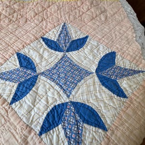 May include: A quilted textile featuring a central diamond-shaped block with a blue and white geometric pattern. The block is surrounded by a soft peach-colored fabric with a grid-like quilted texture and a yellow fabric border trim.