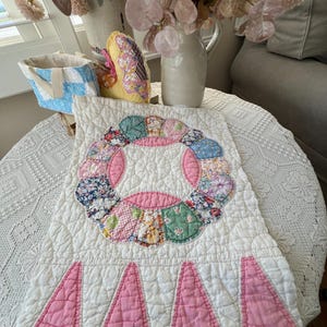 May include: A vintage white quilt with a pink and floral wreath design. The quilt features a scalloped pink border and is displayed on a round table with a white lace tablecloth. A blue and white basket and a yellow pillow are also visible.
