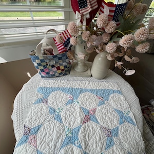 May include: A white and blue quilt with a floral pattern is displayed on a table. A small woven basket and a vase with flowers and American flags are also on the table. A red heart decoration hangs in the background.