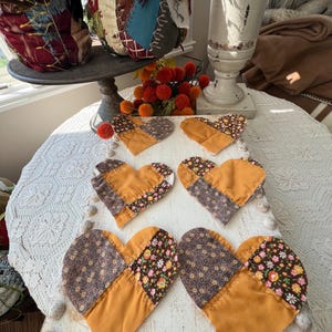 May include: Six heart-shaped fabric coasters arranged in two rows on a white wooden board. Each coaster features a patchwork design combining solid mustard yellow fabric with brown floral patterned fabric, finished with visible white stitching.