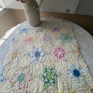May include: A square quilted textile featuring a star-patterned design composed of various floral and plaid fabric patches in shades of pink, blue, green, and yellow, set against a cream-colored background with scalloped edges and visible stitching.