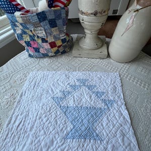 May include: A square white fabric quilt block featuring a central light blue checkered basket design. The piece is quilted with a grid pattern and sits flat, showcasing detailed stitching and a clean, geometric construction on a white surface.