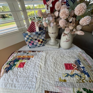 May include: A handmade quilt with a patchwork design featuring red, yellow, blue, and green fabrics. The quilt is placed on a white lace tablecloth. A basket with a patchwork design and American flags are also visible.