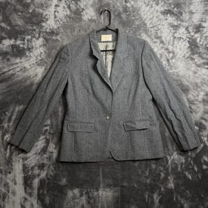 Pode incluir: Um blazer cinza com fecho de um botão, dois bolsos frontais e lapelas entalhadas. O blazer está pendurado em um cabide preto contra um fundo cinza. O blazer é feito de um tecido texturizado.