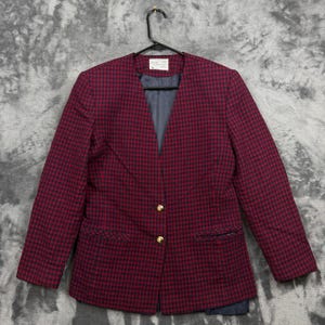 Puede incluir: Una blazer a cuadros burdeos y negro con botones dorados y cuello con solapas. La blazer está colgada de una percha negra sobre un fondo gris. La blazer tiene un bolsillo a cada lado.
