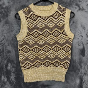 Könnte beinhalten: Eine ärmellose Strickweste mit einem geometrischen Rautenmuster. Die Weste hat eine beige Grundfarbe mit braunen Rautenformen. Der Kragen, die Armausschnitte und der Saum sind einfarbig beige. Die Weste hängt an einem schwarzen Kleiderbügel.