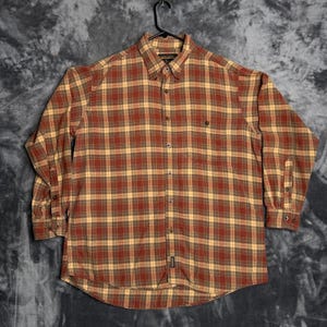 May include: A button-down, long-sleeve plaid shirt in shades of red, brown, and tan. The shirt has a classic collar, a single chest pocket, and buttoned cuffs. The shirt is hanging on a black hanger.