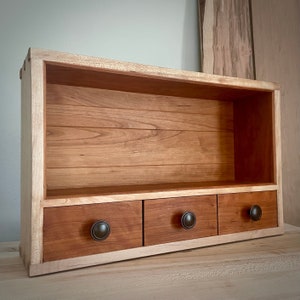 May include: A wooden shelf unit with three drawers. The shelf is made of light wood with a darker wood trim. The drawers have black metal knobs.