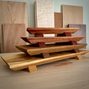 May include: A set of four wooden trays stacked on top of each other. The top two trays are a reddish brown color, while the bottom two trays are a light brown color. The trays are all rectangular and have a slightly curved edge. The trays are made of wood and have a natural finish.
