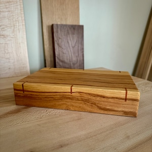 May include: A rectangular wooden cutting board with a light brown finish and red wood inlay. The board has a smooth surface and is made of solid wood.