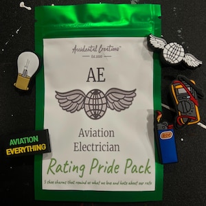 May include: A green pouch with a white label that says "Accidental Creations Est. 2020 AE Aviation Electrician Rating Pride Pack 5 shoe charms that remind us what we love and hate about our rate". The pouch contains five shoe charms: a lightbulb, a pair of wings with a globe, a multimeter, a lighter, and a black and green charm that says "AVIATION EVERYTHING".