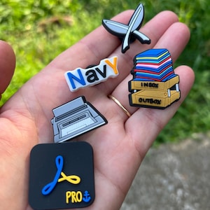 May include: Five colorful shoe charms featuring a typewriter, a stack of papers with an inbox and outbox label, a navy blue word charm, a yellow and blue swirl design with the word "PRO" and an anchor, and a white and gray crossed feather design.