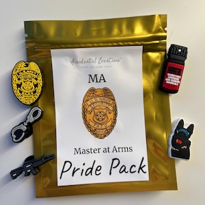 Może przedstawiać: Złota saszetka z zamkiem błyskawicznym z białą etykietą, na której widnieje napis "Accidental Creations" i "Est 2020". Etykieta zawiera również napis "MA" i "Master at Arms Pride Pack". Saszetka zawiera różne czarne i czerwone gumowe breloczki, w tym odznakę z napisem "U.S. Navy Security FAFO 1973", kajdanki, pistolet, butelkę gazu pieprzowego i czarnego kota.