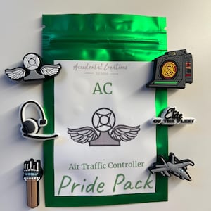 May include: A green pouch with a white label that says "Accidental Creations Est. 2020 AC Air Traffic Controller Pride Pack". The pouch contains six colorful charms, including a black and yellow radar, a white radio with wings, a black headset, a white and gray airplane, a black and white "Elite of the Fleet" charm, and a brown and white control tower.
