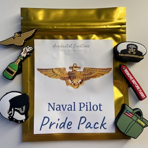 May include: A gold zippered pouch containing a set of 7 colorful rubber shoe charms. The charms include a champagne bottle, a pilot's wings, a naval officer's hat, a "Remove Before Flight" tag, a pilot's helmet, a US Navy bag, and a gold pilot's wings with an anchor. The pouch has a white label with the text "Accidental Creations Est. 2020 Naval Pilot Pride Pack".