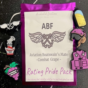 Puede incluir: Una bolsa morada con una etiqueta blanca que dice "Accidental Creations Est 2010" y "ABF Aviation Boatswain's Mate -Combat Grape- Rating Pride Pack 5 shoe charms that remind us what we love and hate about our rate". La bolsa contiene cinco amuletos para zapatos: un ancla blanca con alas, una caricatura roja y blanca de un motor de barco, una uva morada con hojas verdes, un casco morado con una visera negra y un chaleco salvavidas morado.
