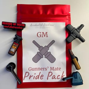 May include: A red pouch with the text "Accidental Creations Est. 2020 GM Gunners' Mate Pride Pack" and a black and white image of two crossed wrenches. The pouch contains a variety of small black, brown, and blue rubber charms.