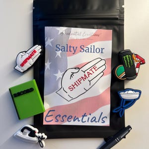 May include: A black zippered pouch with a white label that says "Salty Sailor Essentials" with an American flag design and a hand with the word "SHIPMATE" written on it. The pouch contains a variety of colourful rubber charms, including a green record, a blue ship, a white iron, a black pen, and a green monster energy drink can with a pack of cigarettes.