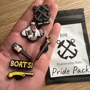 Puede incluir: Un conjunto de seis abalorios para zapatos coloridos que representan un mono con un sombrero de marinero, un pincel, un cañón, una espada y dos anclas. Los abalorios están empaquetados en una bolsa negra con el texto "Boatswain's Mate Pride Pack".