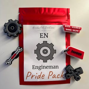 Op de afbeelding: Een rode hersluitbare zak met een wit label waarop staat "Accidental Creations Est. 2020 EN" en "Engineman Pride Pack". De zak bevat vijf kleine, kleurrijke rubberen bedels met verschillende ontwerpen, waaronder een moersleutel, een tandwiel, een gevaarsbord en een zuiger.