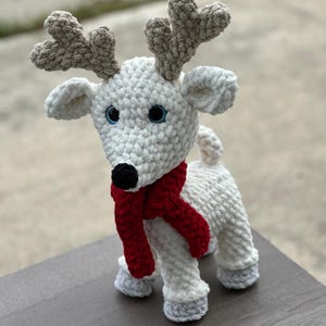 May include: A handmade crochet reindeer with a white body, gray antlers, and gray hooves. It has a black nose, blue eyes, and a red scarf. The plush toy is standing on a wooden surface.