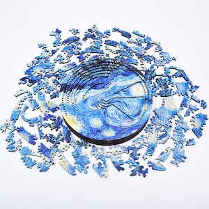 Puede incluir: Un rompecabezas de madera circular con la pintura "La noche estrellada" de Van Gogh. La imagen central está completa, rodeada de numerosas piezas azules dispersas. El diseño y la paleta de colores crean una escena visual atractiva.