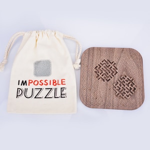 Puede incluir: Un rompecabezas de madera con un diseño de laberinto complejo y dos piezas extraíbles se encuentra junto a una bolsa con cordón. La bolsa es de color crema y tiene la inscripción "IMPOSSIBLE PUZZLE" en negro y rojo.