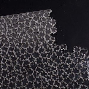 Puede incluir: Un rompecabezas claro e interconectado con un dise&ntilde;o geom&eacute;trico complejo. Las piezas del rompecabezas son transparentes y est&aacute;n dispuestas en un patr&oacute;n repetitivo, con una secci&oacute;n faltante, revelando el fondo negro.
