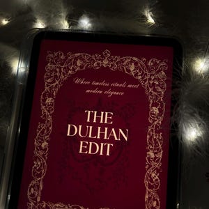 Puede incluir: Una tableta digital muestra una portada burdeos con detalles dorados ornamentados y el texto "THE DULHAN EDIT". La portada tambi&eacute;n incluye el texto "Where timeless rituals meet modern elegance" y "By Attar & Amber". La tableta est&aacute; rodeada de plumas blancas y luces de hadas.
