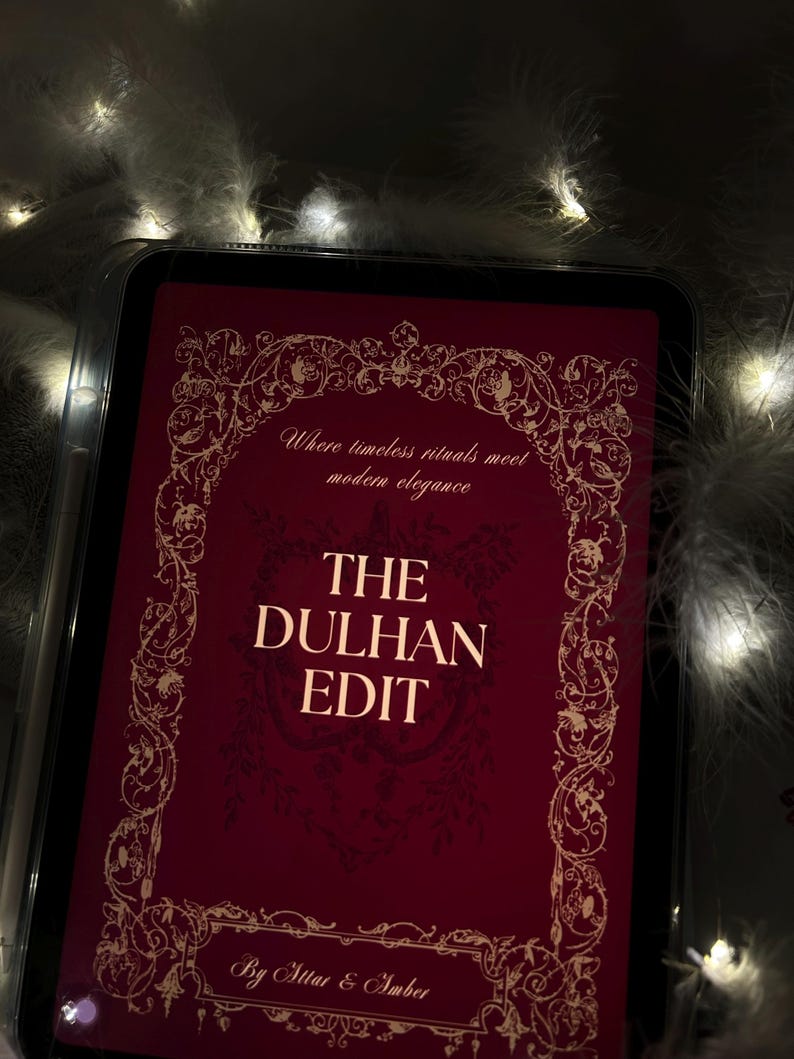 Puede incluir: Una tableta digital muestra una portada burdeos con detalles dorados ornamentados y el texto "THE DULHAN EDIT". La portada tambi&eacute;n incluye el texto "Where timeless rituals meet modern elegance" y "By Attar & Amber". La tableta est&aacute; rodeada de plumas blancas y luces de hadas.