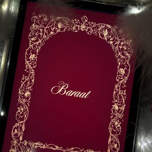 Puede incluir: Una pantalla de tableta de color burdeos muestra la palabra "Baraat" en una elegante escritura, enmarcada por dise&ntilde;os florales dorados. El texto "Attar & Amber" est&aacute; debajo. La imagen est&aacute; rodeada de suaves plumas blancas y luces de hadas.