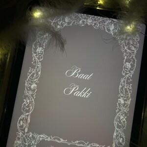 Puede incluir: Una tableta digital muestra una pantalla gris claro con un borde floral blanco y texto. El texto dice "Baat Pakki" y "Attar & Amber". La imagen est&aacute; enmarcada por un borde negro e iluminada por luces suaves y c&aacute;lidas y plumas blancas.
